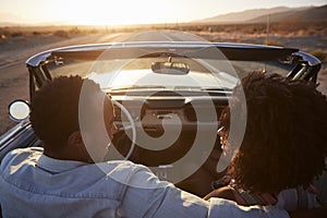 Rear View Of Couple On Road Trip Driving Classic Convertible Car Towards Sunset