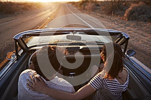 Rear View Of Couple On Road Trip Driving Classic Convertible Car Towards Sunset