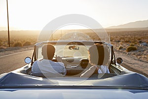 Rear View Of Couple On Road Trip Driving Classic Convertible Car Towards Sunset