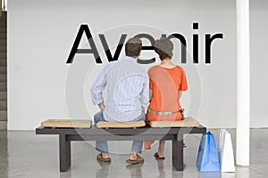 Rear view of couple seated on bench reading French text Avenir (future) on wall