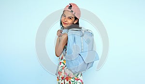 Rear view of a cute kid smiling wearing colorful dress, pink cap, and blue backpack looking at the camera through her arm posing