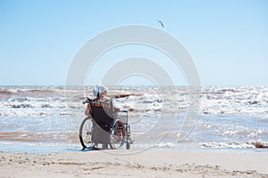 Rear view of a disabled woman sitting on a wheelchair on the beach on a sunny day. She looks at the ocean