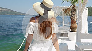 Rear view of elegant woman in hat and long dress relaxing by walking at the wooden sea pier on a sunny summer day