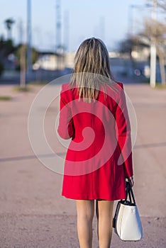 Rear view of an elegant woman wearing red jacket, skirt and holding a white handbag while walking in the street in a sunny day