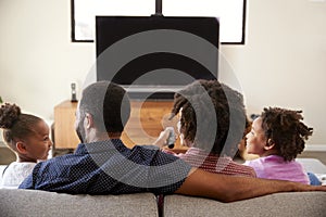 Rear View Of Family With Children Sitting On Sofa Watching TV Together