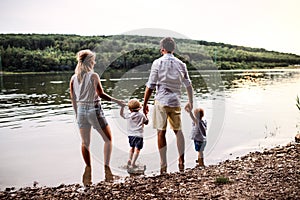 A rear view of family with two toddler children outdoors by the river in summer.