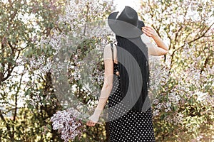 Rear view on fashionable woman with long hair in stylish dress in fashionable hat with bouquet lilac flowers near flowering tree