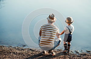 A rear view of father with a small toddler son spending time outdoors by a lake.