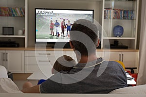 Rear view of father and son sitting at home together watching sport event on tv