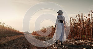 Rear view: A female farmer in a dress and hat walks along the fields of ripe corn.