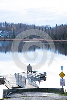 Rear view fisherman sitting on small backpacking chair on fishing dock with barrier edge protection, autumn forest with row of