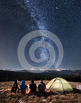 Rear view of four people sitting together beside camp and tent under beautiful night sky full of stars and milky way