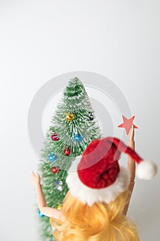 Rear view of girl doll in Santa hat holding star decoration next to fir tree and preparing for Christmas home decoration minimal