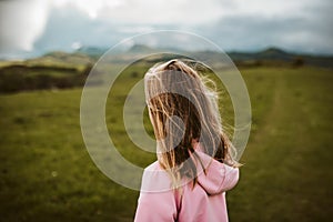 Rear view of girl on walk in spring nature, rain clouds on sky. Wearing rain jacket and rubber rain boots. Spring or