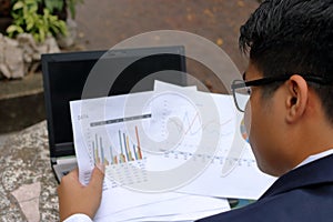 Rear view of handsome businessman looking documents charts and thinking about his job in the park outdoors