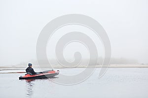 Rear view of handsome man sitting in canoe and looking in distance, guy wearing black jacket and cap, foggy autumn morning,