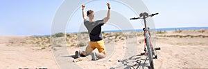 Rear view of happy excited winner traveler kneel down with raised hands, next to him is bicycle