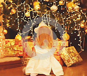 Rear view happy little child sitting and dreaming near christmas tree with garland lights and gift boxes home
