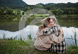 Rear view of happy senior mother embracing with adult daughter when sitting by lake outdoors in nature