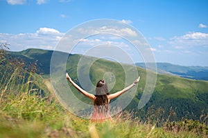 Rear view of a happy woman hiker in red dress sitting on grassy hill on a windy day in summer mountains with outstretched arms