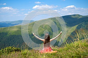 Rear view of a happy woman hiker in red dress sitting on grassy hill on a windy day in summer mountains with outstretched arms