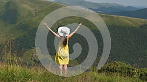 Rear view of a happy woman hiker in yellow dress standing on grassy hill on a windy day in summer mountains with