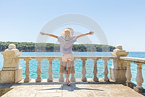 Rear view of happy woman on vacation, wearing straw summer hat ,standing on luxury elegant old stone balcony of coastal