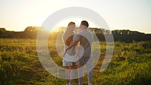 Rear view of happy young couple in love taking each other hand and walking across field towards sunset on beautiful