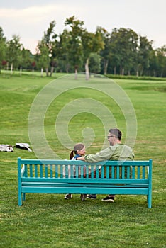 Rear view of happy young father and his cute little daughter talking while sitting together on the bench, spending time