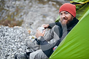 Rear view of happy young male drinking hot beverage in mountains. Traveler man with beard wearing red hat, sitting near to camping