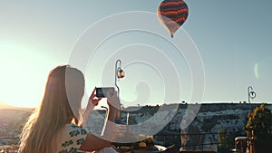 Rear view happy young tourist woman using smartphone to capture video of incredible hot air balloon in the sky.