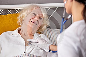 rear view on helpful female nurse checking sounds in heart and lungs of sick senior woman lying on bed