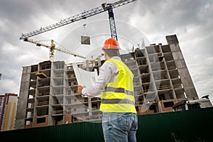 Rear view image of construction engineer in green safety vest and red hardhat controlling construction of new building