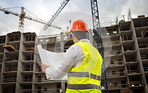 Rear view image of construction engineer looking at blueprints and working cranes on building site