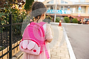 Rear view image of cute little girl preschooler walking outdoor with pink backpack against blurred buildings. Happy kid toodler