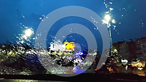rear view from inside mirror rain water drops falling on car window windshield glass with blurred traffic lights and