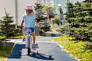 Rear view of little child in summer hat riding bike outdoors in city park