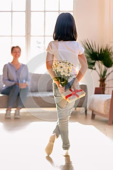 Rear view little daughter holding flowers and gift, congratulate mother