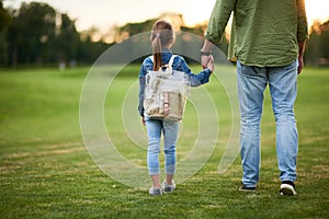 Rear view of little girl with backpack holding father`s hand, while spending time with her daddy in the green park on a