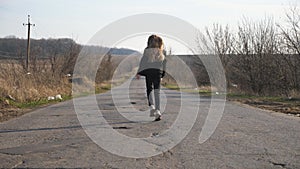 Rear view of little girl in jacket walks along empty road and dancing funny at sunny day. Small female child with long