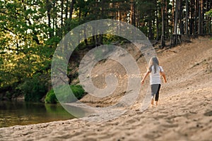 Rear view of little girl with long hair run on sandy beach near river in forest. Female child actively hike in nature.