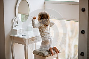 Rear view of little girl in pajamas sitting in front of dressing table with mirror