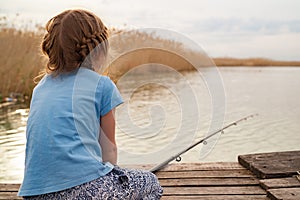 rear view. little girl sits on a wooden bridge on a river with fishing rod.