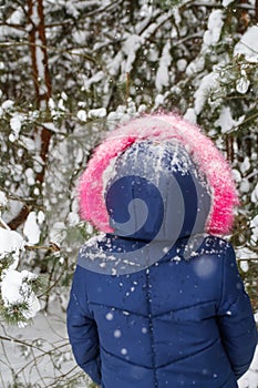 Rear view of little happy girl in pink warm fur hood walking outside on nature winter snowy forest background. Pretty