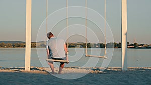 Rear view of a lonely lonely man swinging on a swing alone on the beach. Male loneliness on the shore of the lake.