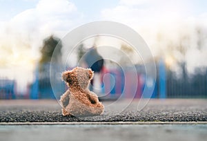 Rear view Lonely Teddy bear doll sitting alone on footpath with blurry kid playing playground in retro filter, Back view Lost