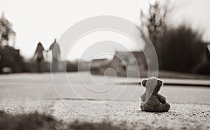 Rear view Lonely Teddy bear doll sitting alone on hailstones with blurry couple walking on footpath.lost brown bear toy looking