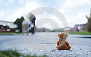 Rear view Lonely Teddy bear doll sitting alone on hailstones with blurry father pushing stroller walking on footpath.lost brown