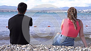 Rear view of loving couple sitting on stony shore in front of the sea. Media. Honeymoon concept, man and woman in love
