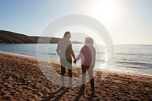 Rear View Of Loving Retired Couple Holding Hands Looking Out To Sea On Winter Beach Vacation